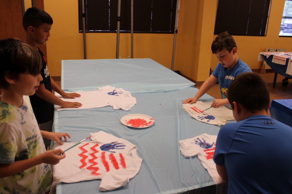 Group of campers at South Cape Community Center summer camp stand around a long table painting white T-shirts with red and blue patriotic designs for 4th of July.